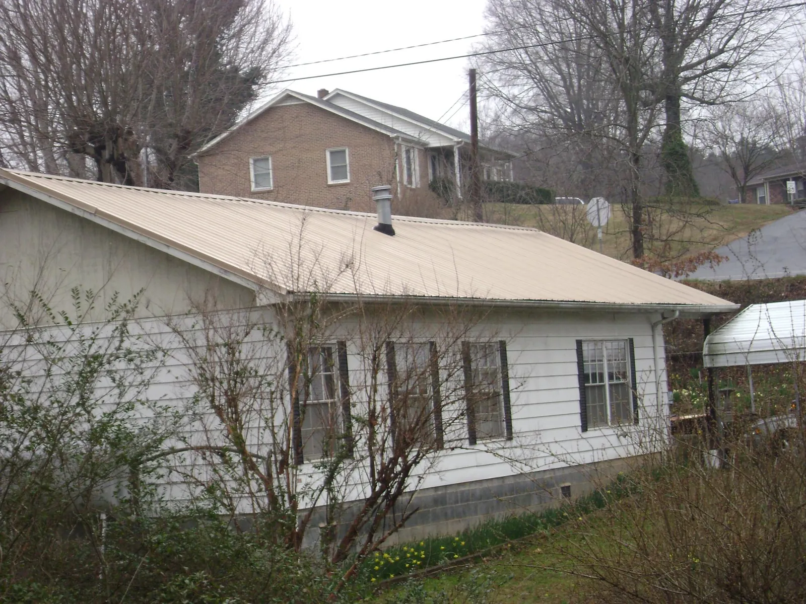 Tan metal roof on a white sided house set into a hillside — Commercial Roofing by Roofing Brothers in Brookline